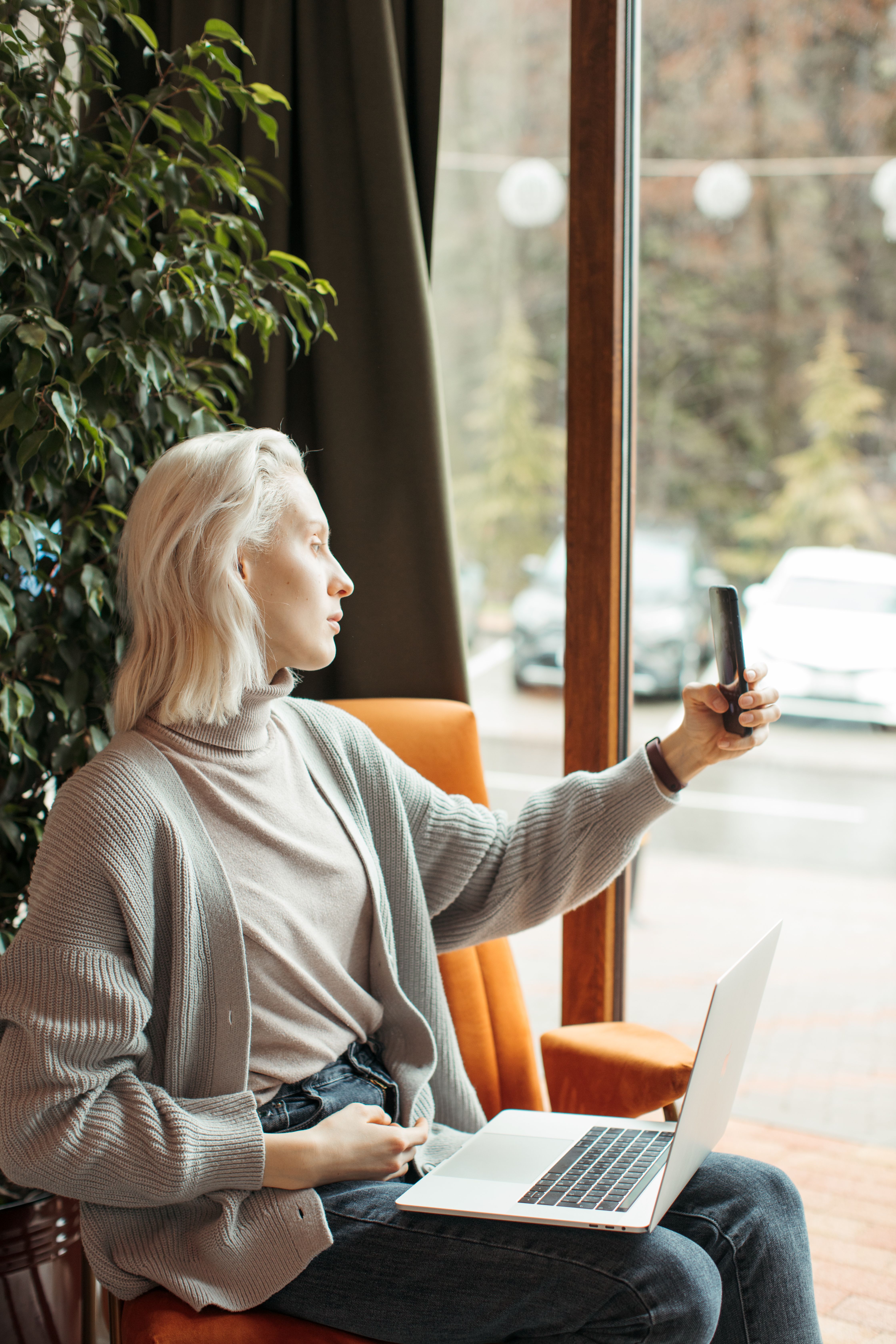 Blonde woman takes a selfie with her smartphone while sitting indoors.
