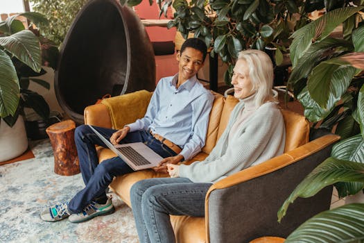 Two colleagues smiling and collaborating on a laptop in a cozy, plant-filled office setting.