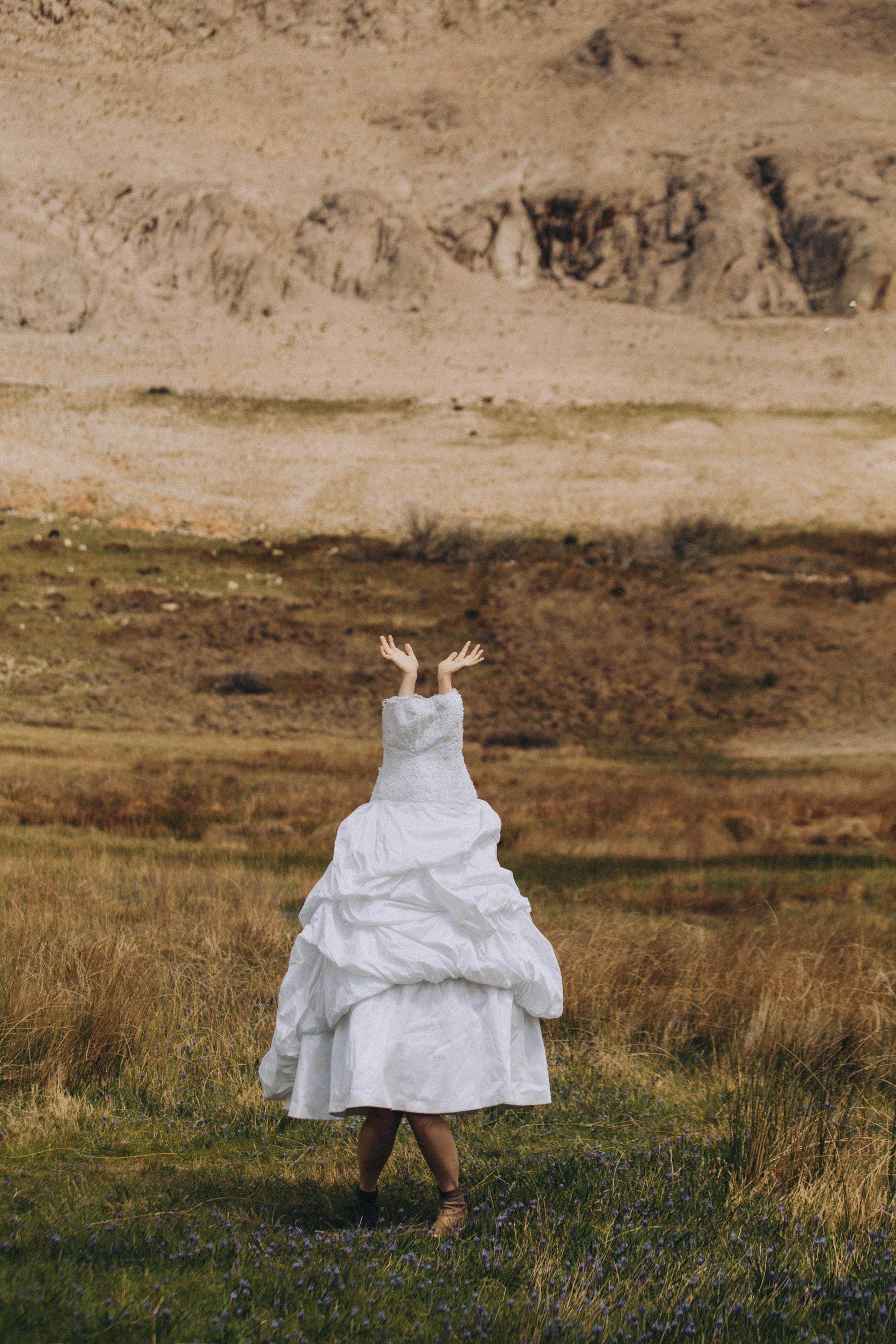 A woman in a white wedding dress reaches up in a grassy field against a rugged mountain backdrop.