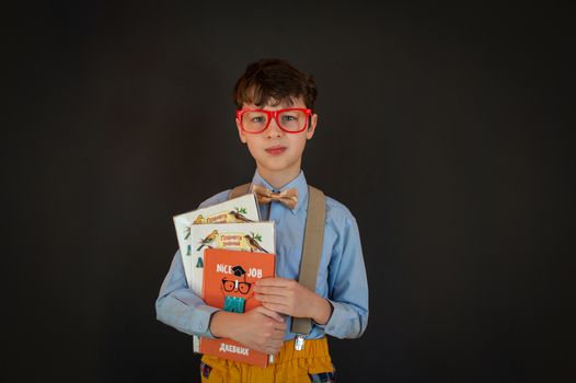 Smart little boy in school uniform with glasses and suspenders holding books while looking at camera and standing against black wall
