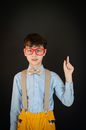 Nerd schoolboy in red eyeglasses blue shirt with bow tie and yellow trousers with suspenders pointing index finger up