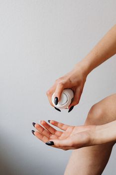 Close-up of a woman's hands with black nail polish applying skincare cream against a light background.