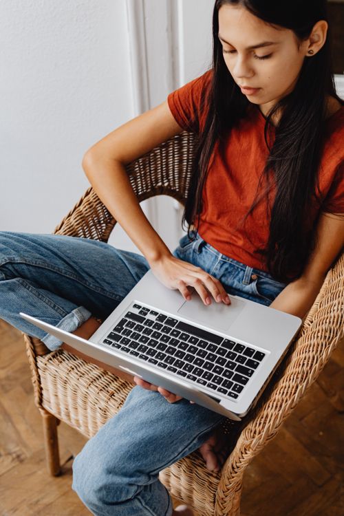 Free Focused young woman using a laptop while sitting comfortably in a wicker chair indoors. Stock Photo