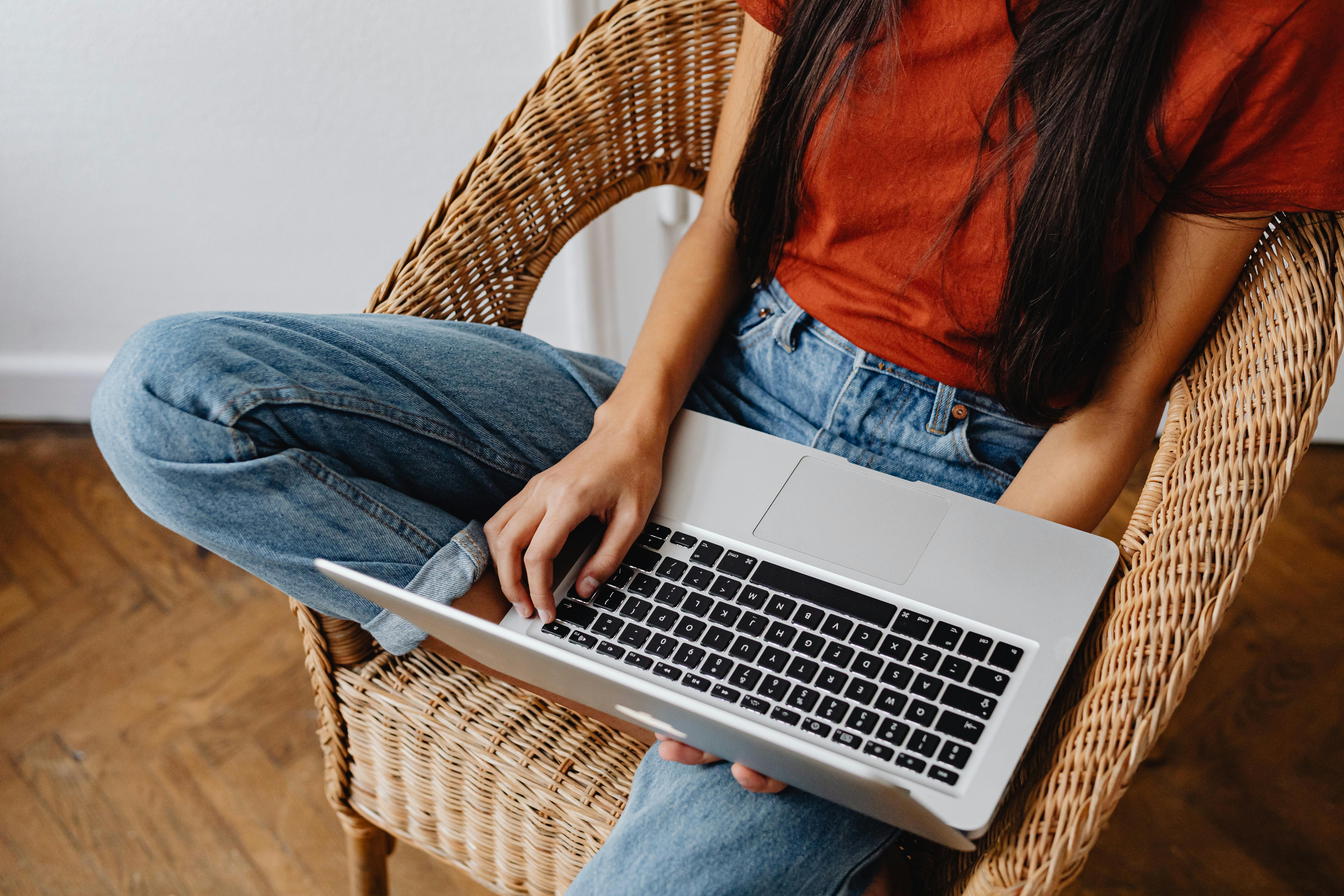 Young woman comfortably using a laptop while sitting on a woven chair indoors.