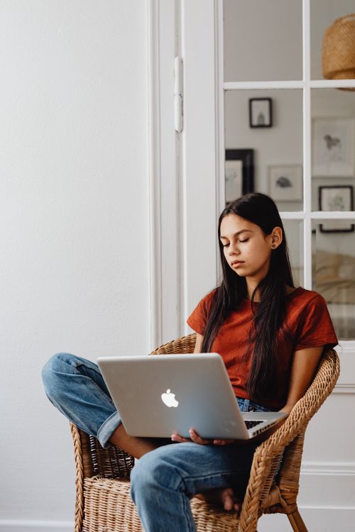Free Young woman sitting in a wicker chair using a silver laptop, relaxing indoors near a window. Stock Photo