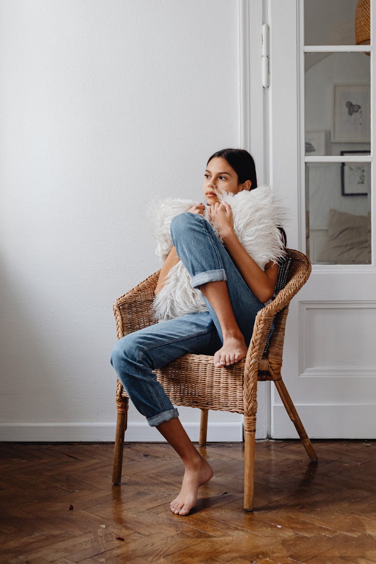 Girl Sitting On Brown Woven Chair Hugging White Furry Pillow