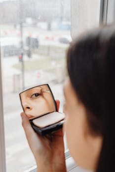 Close-up of a woman applying eye makeup using a compact mirror near a window.
