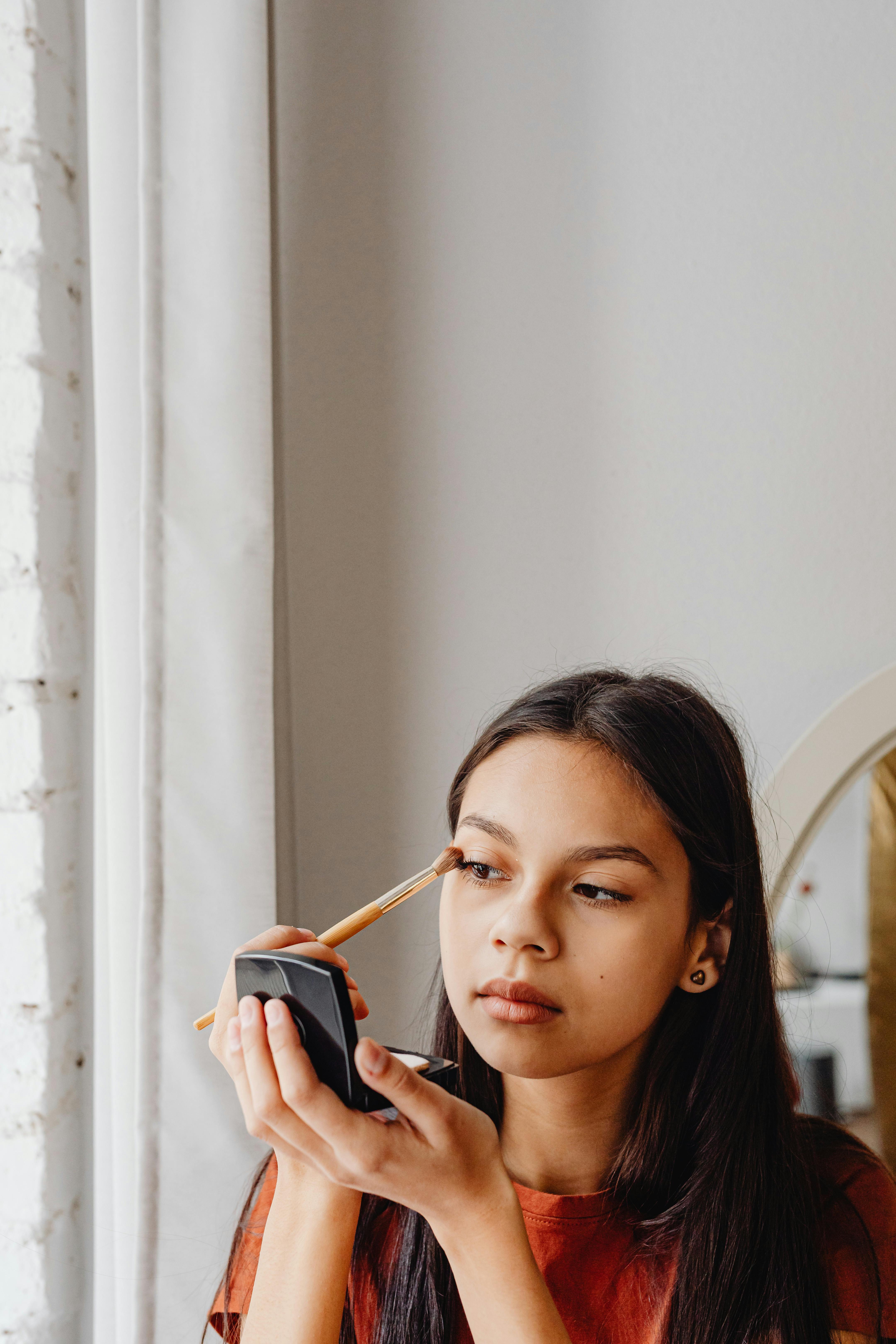 Young woman applying eye makeup using a compact mirror in a cozy indoor setting.