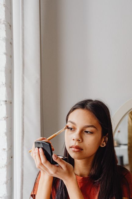 Young woman applying eye makeup using a compact mirror in a cozy indoor setting.