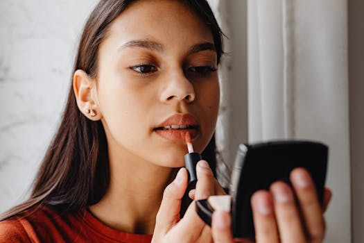 A young woman applies lipstick while looking in a handheld mirror.