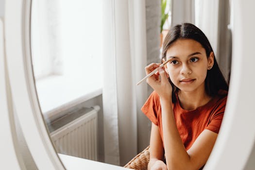 Young woman using a pencil for eyebrow makeup in front of a mirror in a cozy room.
