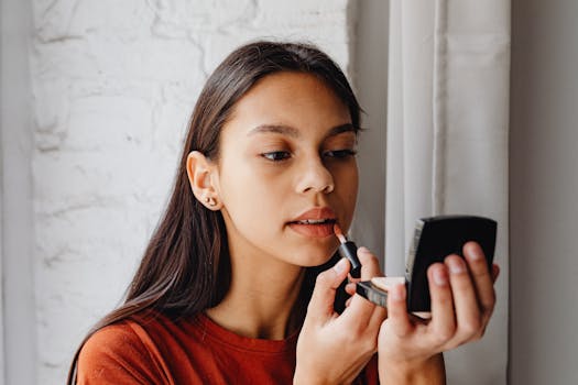 Young woman applying lip tint using a compact mirror indoors by a window.
