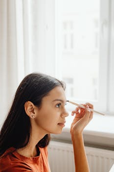 A young woman skillfully applies makeup to her eyebrows by a bright window indoors.
