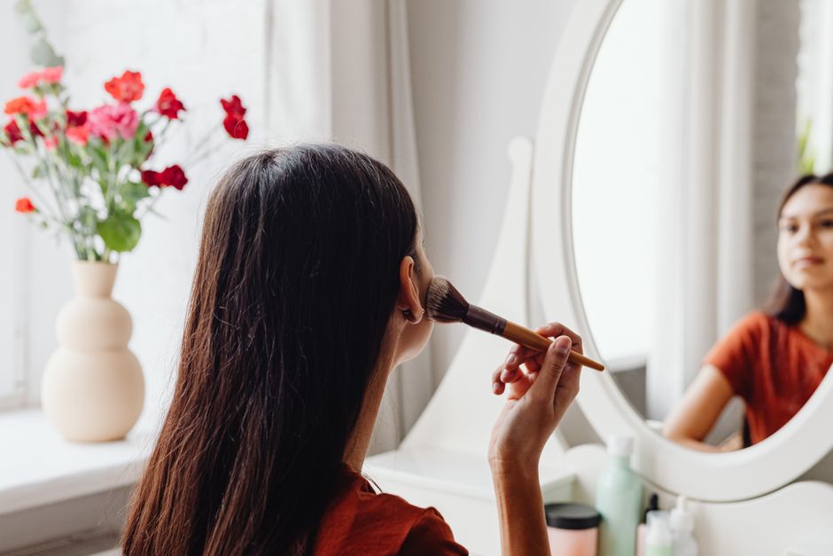A woman applying makeup with a brush while facing a mirror indoors.