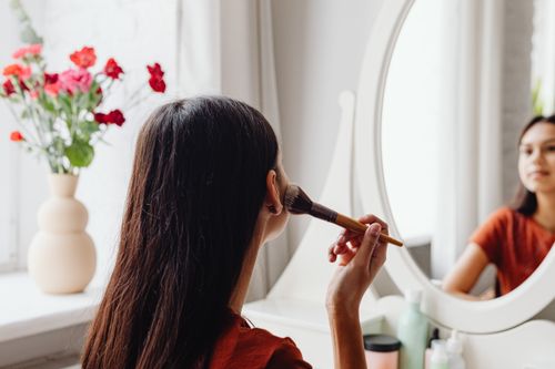 Free A woman applying makeup with a brush while facing a mirror indoors. Stock Photo