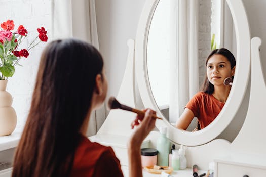 A young woman applies makeup with a brush in front of a stylish vanity mirror, adding elegance to her morning routine.