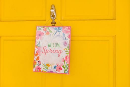 A colorful 'Welcome Spring' sign with floral motifs hangs on a vibrant yellow door, symbolizing renewal.