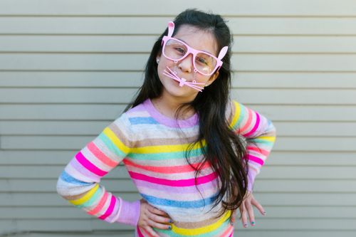 Free A young girl wearing pink bunny glasses and a colorful striped sweater poses playfully outdoors. Stock Photo