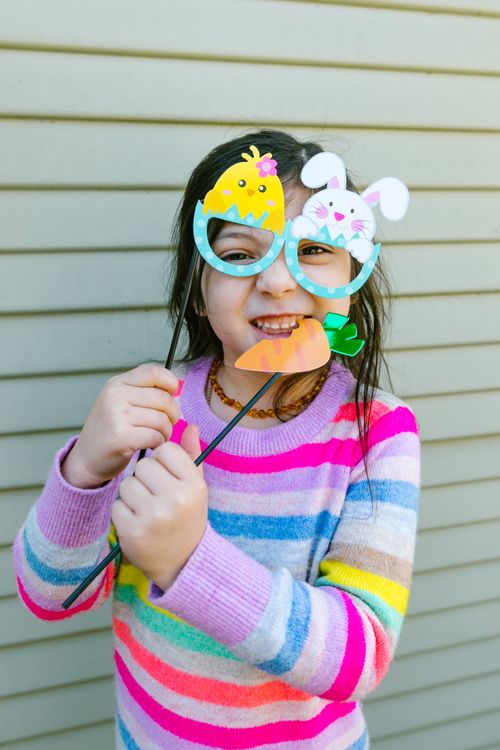 Free A young girl in a colorful striped sweater poses with Easter-themed glasses, smiling joyfully. Stock Photo