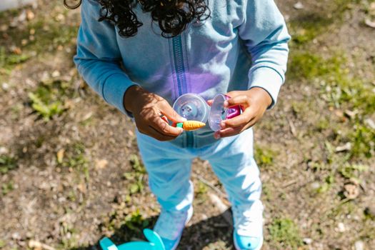 A child in blue clothes holds an opened Easter egg with a toy carrot, enjoying an Easter egg hunt outdoors.