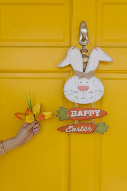 Free Charming Easter bunny decoration on a vibrant yellow door with a hand holding flowers. Stock Photo