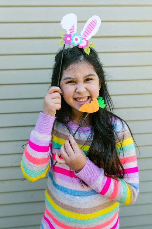 Free Smiling child holding bunny ears and a carrot decoration, in a colorful sweater against a striped backdrop. Stock Photo