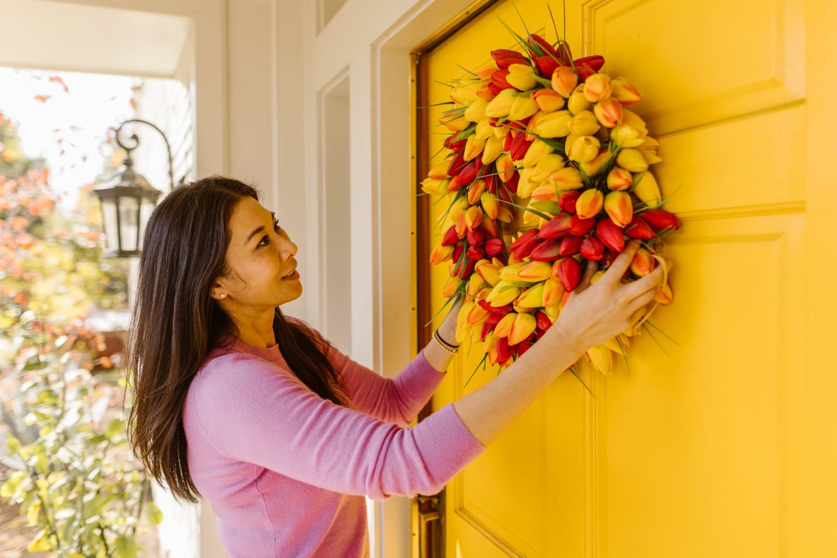 A woman hangs a vibrant floral wreath on a bright yellow door, symbolizing spring decor. Stock Photo.