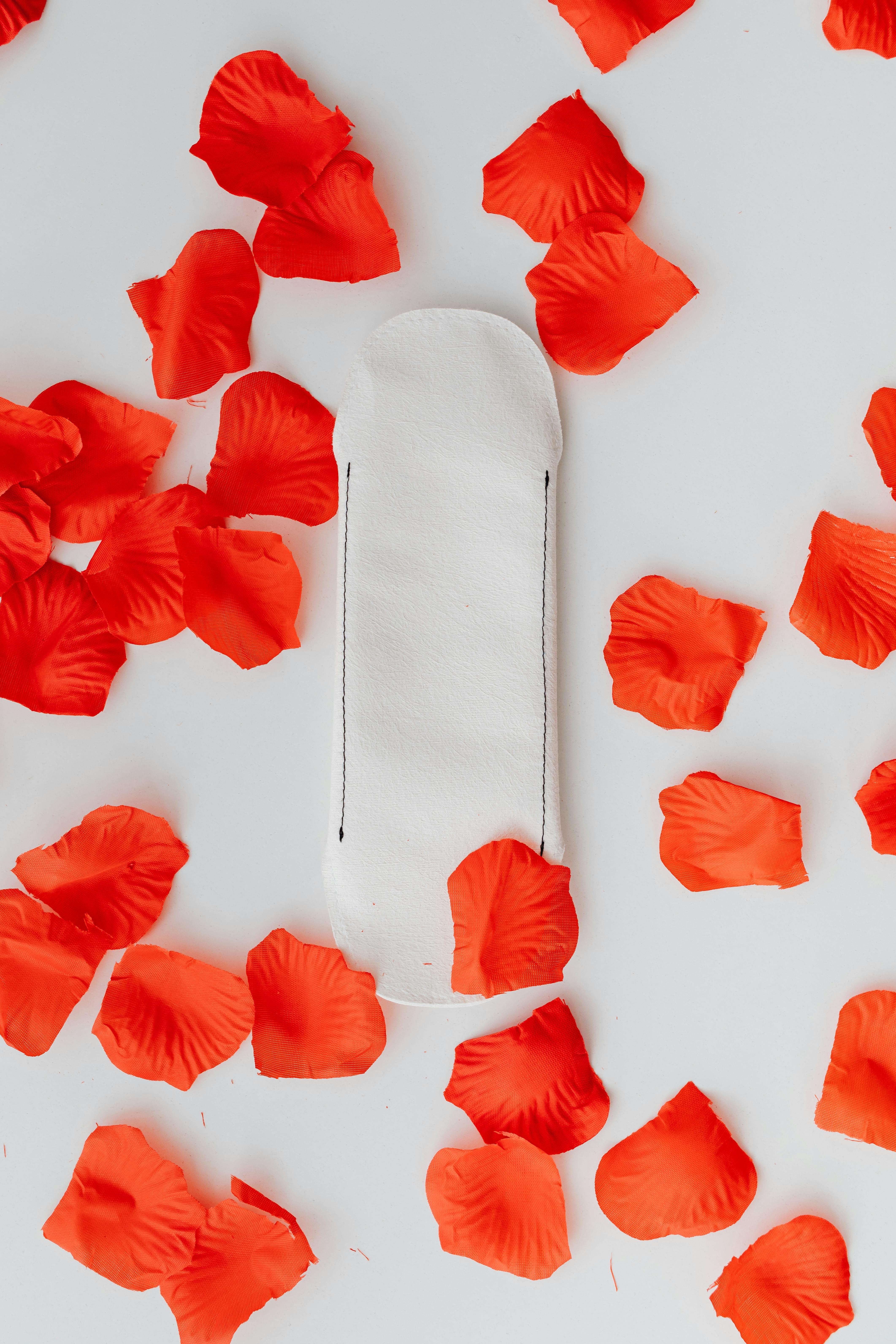 A close-up of a sanitary pad surrounded by red petals on a white background, representing feminine hygiene.