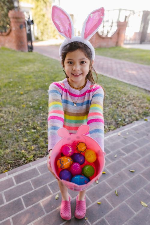 Free A smiling child with bunny ears holds a colorful basket of Easter eggs outdoors. Stock Photo