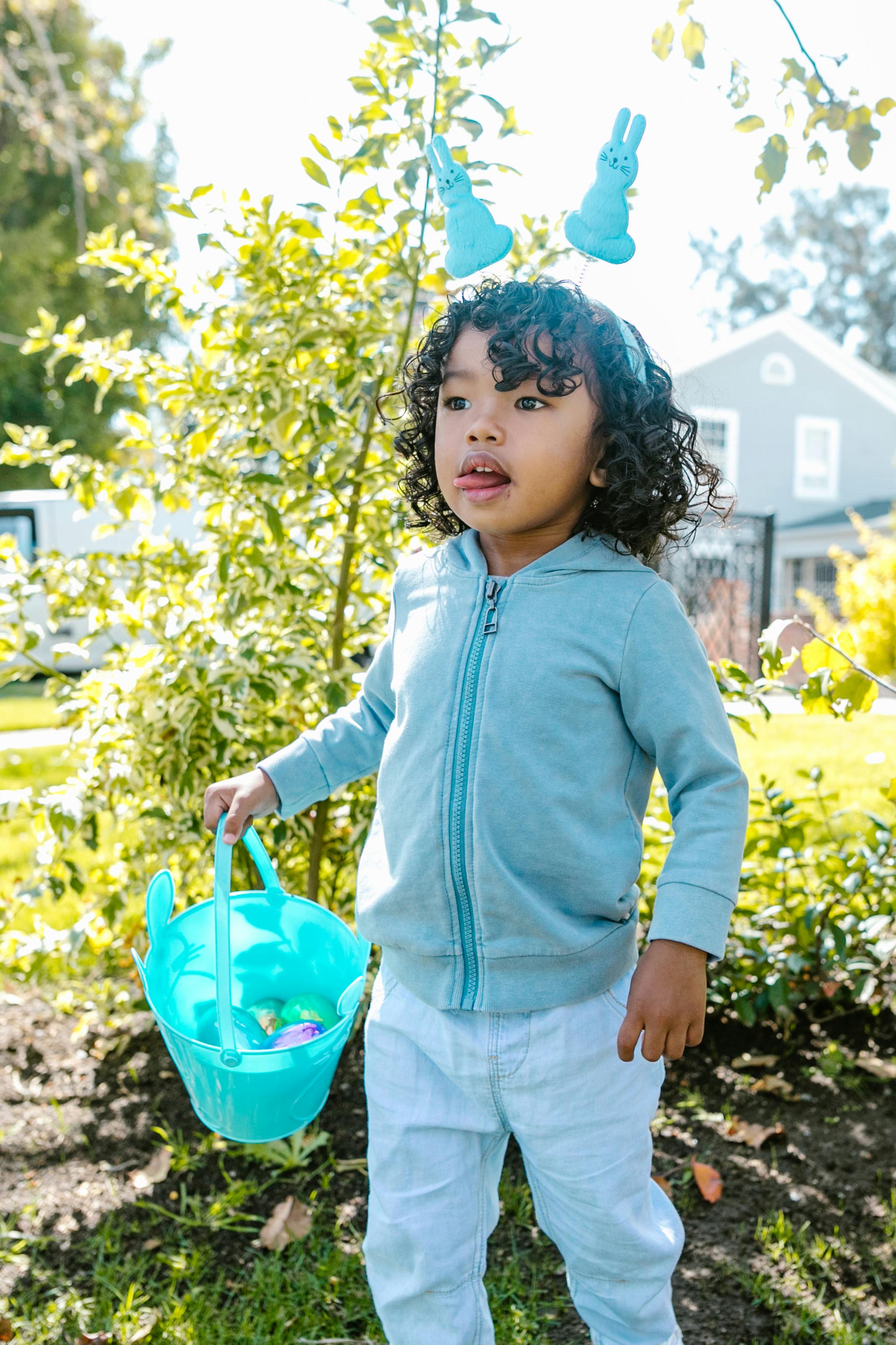 A child with bunny ears holds a bucket filled with Easter eggs during a sunny outdoor Easter egg hunt.