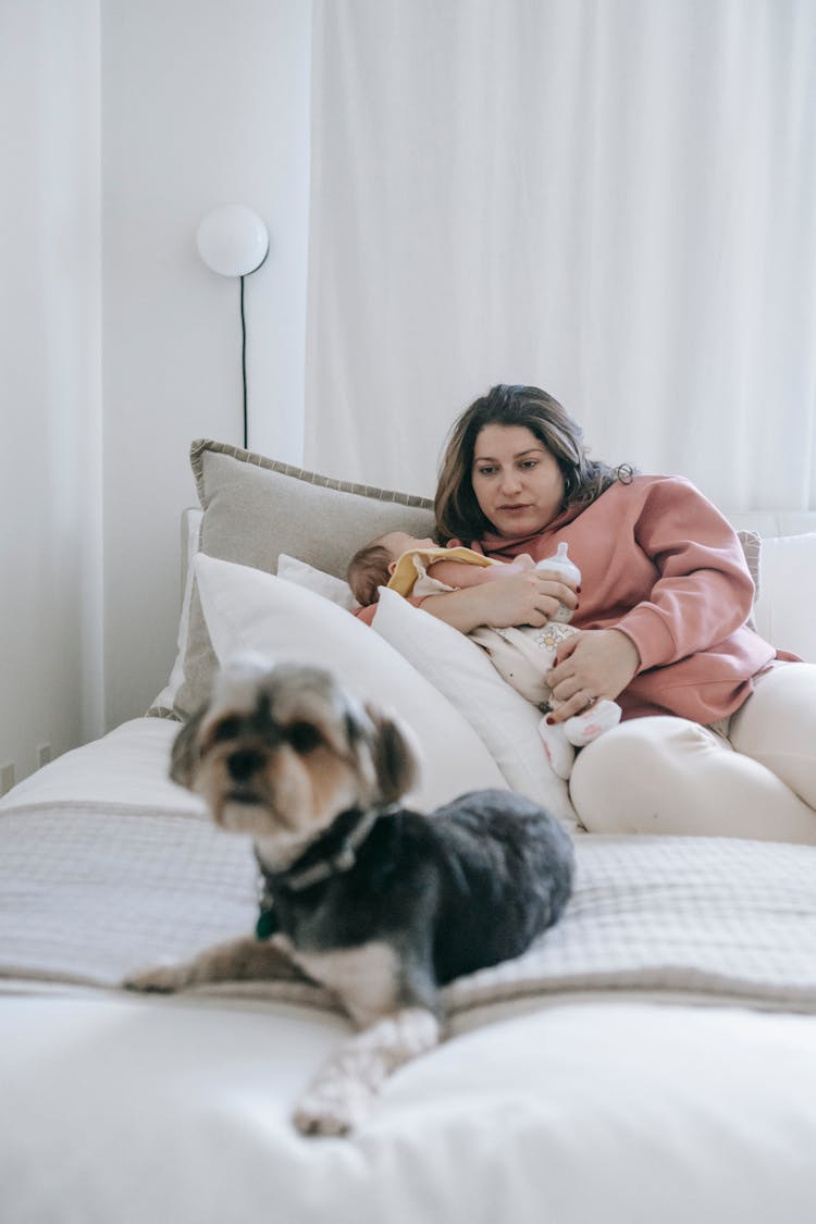 Woman Carrying A Baby Lying On A Bed With A Dog
