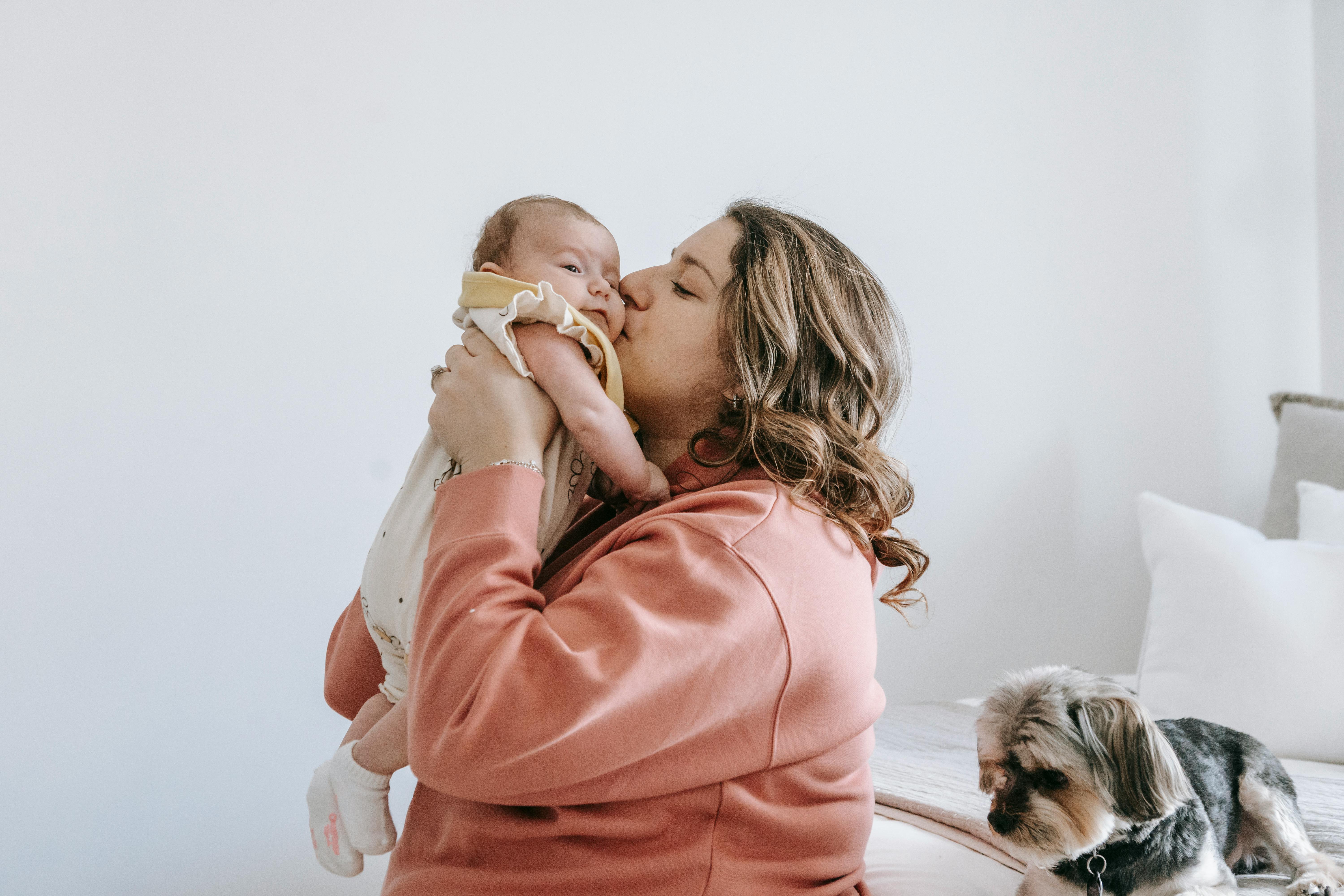 Side view of happy woman kiss cute little baby while in bright room with puppy sitting on bed and