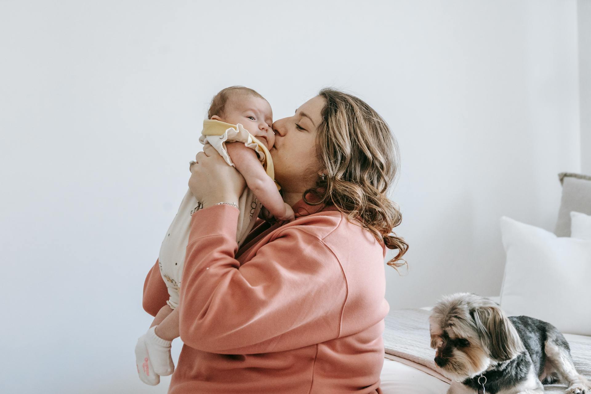 Zicht van de zijkant van gelukkige vrouw kus schattig baby terwijl in lichte kamer met puppy zitten op bed en