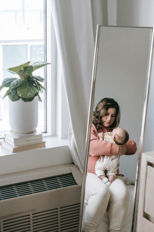 Free In reflection of mirror young mother sitting on bed in right room with adorable little baby in arms Stock Photo