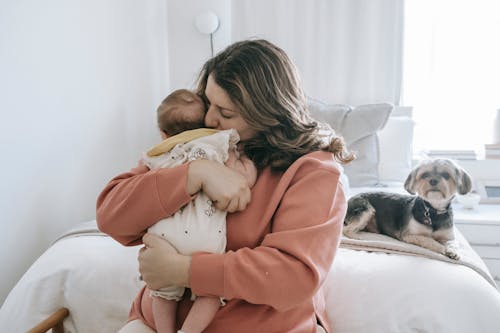 Free Beautiful female hugging little baby sitting on bed while their puppy looking at them Stock Photo