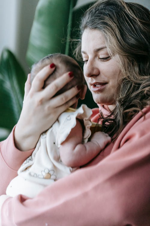 Free Side view of young mother in casual clothes with cute little girl in hands sitting at home Stock Photo