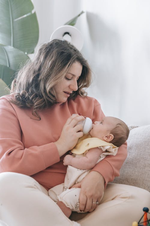 Free Caring mother feeding cute infant baby with bottle while sitting on comfortable sofa in light room with potted plant at home Stock Photo
