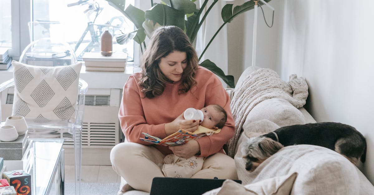 Caring mother feeding baby with bottle while sitting on sofa with laptop in modern room with dog and potted plant