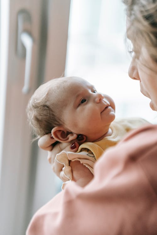 Side view of crop mother with adorable little newborn baby looking at each other while standing near window in room