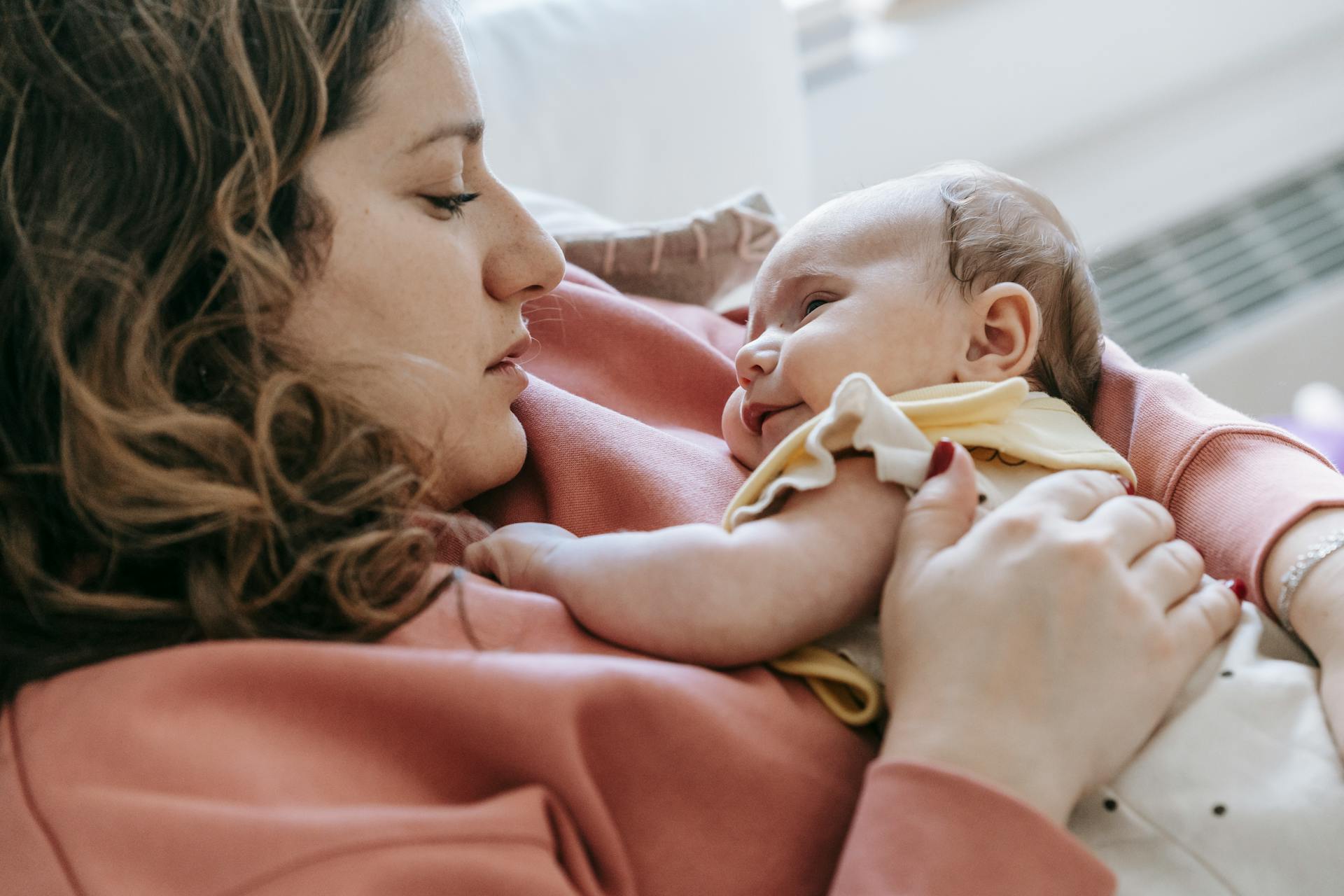 Side view of caring mother with cute newborn baby in arms looking at each other while sitting on couch in room