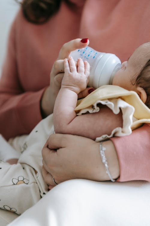 Free Crop unrecognizable mother in casual wear feeding crop adorable infant with bottle in arms while sitting in light room at home Stock Photo