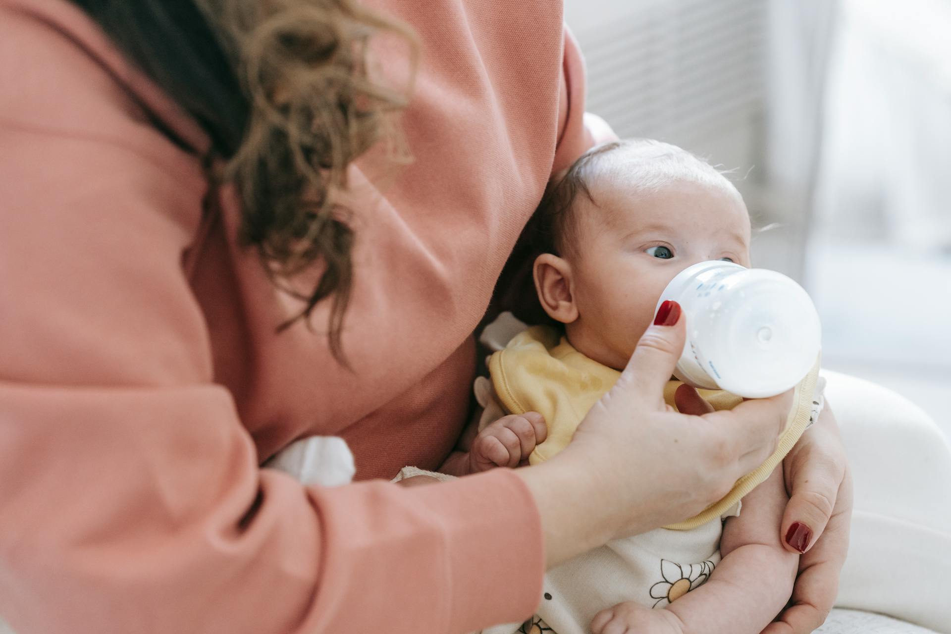 Crop anonymous mother in casual clothes feeding adorable newborn baby with bottle while sitting on sofa in light room at home