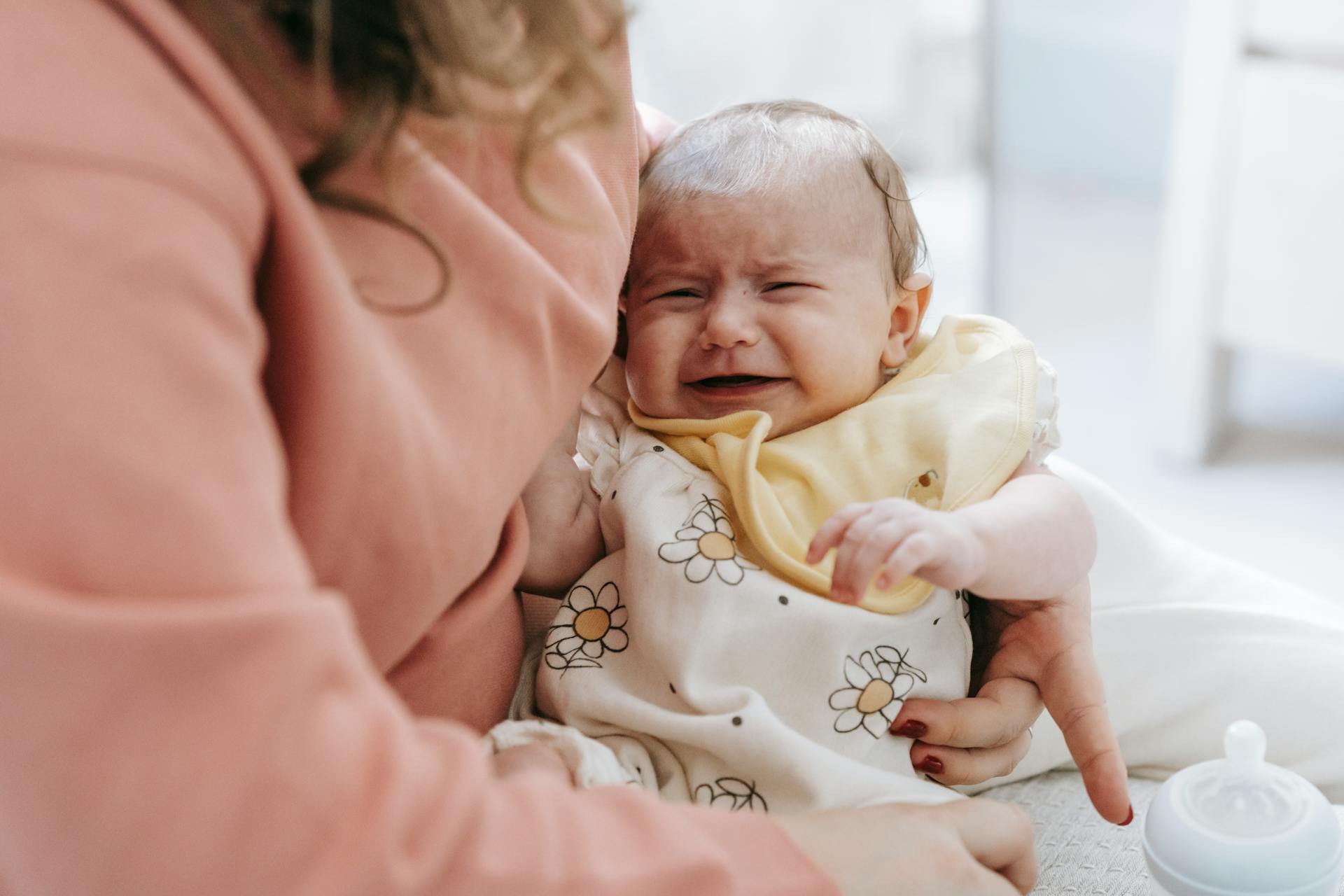 Crop anonymous caring mother with crying newborn baby in arms sitting on couch in light room with feeding bottle at home