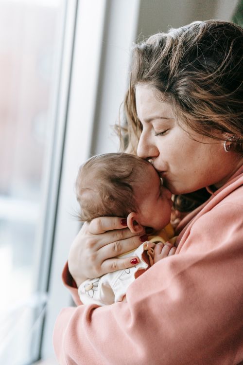 Free Side view of young woman with long hair hugging and kissing newborn baby with closed eyes while standing near window at home Stock Photo