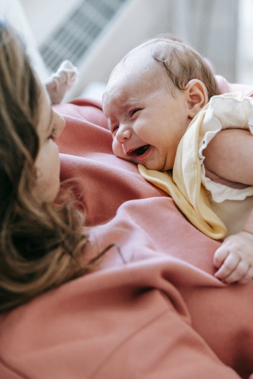 Free From above of adorable newborn baby crying in hands of crop young mother sitting on sofa at home Stock Photo