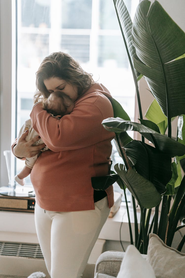 Young Mother Hugging Baby With Closed Eyes Near Window