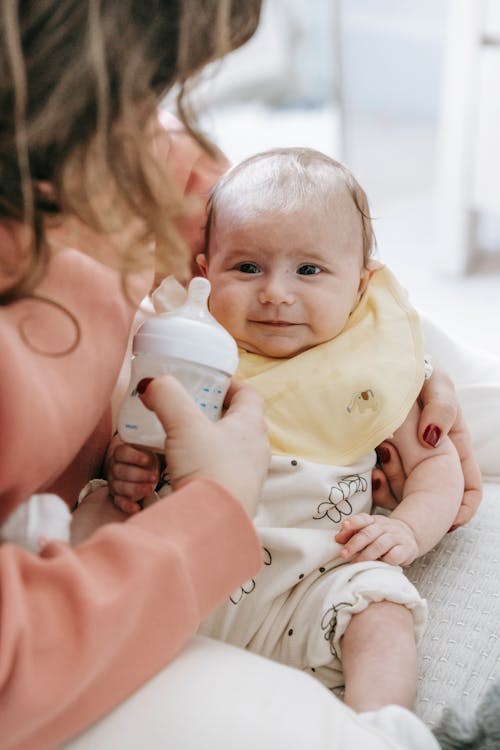 Free Crop unrecognizable female with wavy hair in casual clothes hugging adorable newborn during feeding from bottle on sofa at home Stock Photo