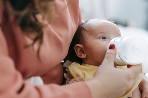 Crop faceless mother in casual clothes embracing and feeding cute newborn baby with milk from bottle in daylight at home