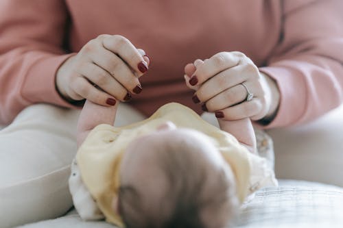 Crop faceless mom holding hands of cute baby lying on bed after changing diaper in daylight