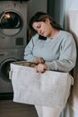 Focused female with bag of clothes having phone conversation while standing in washroom near modern washing machines during housework routine
